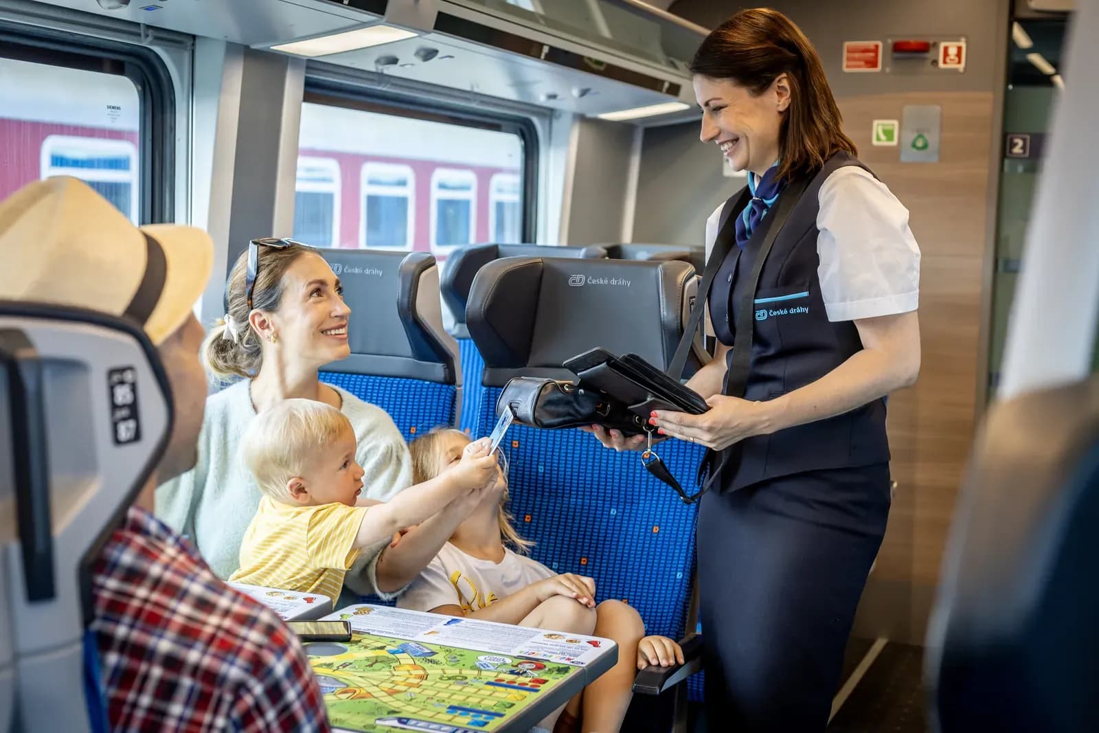 Smiling České dráhy conductor holding a portable payment terminal as a small boy hands her a card at a family’s seat