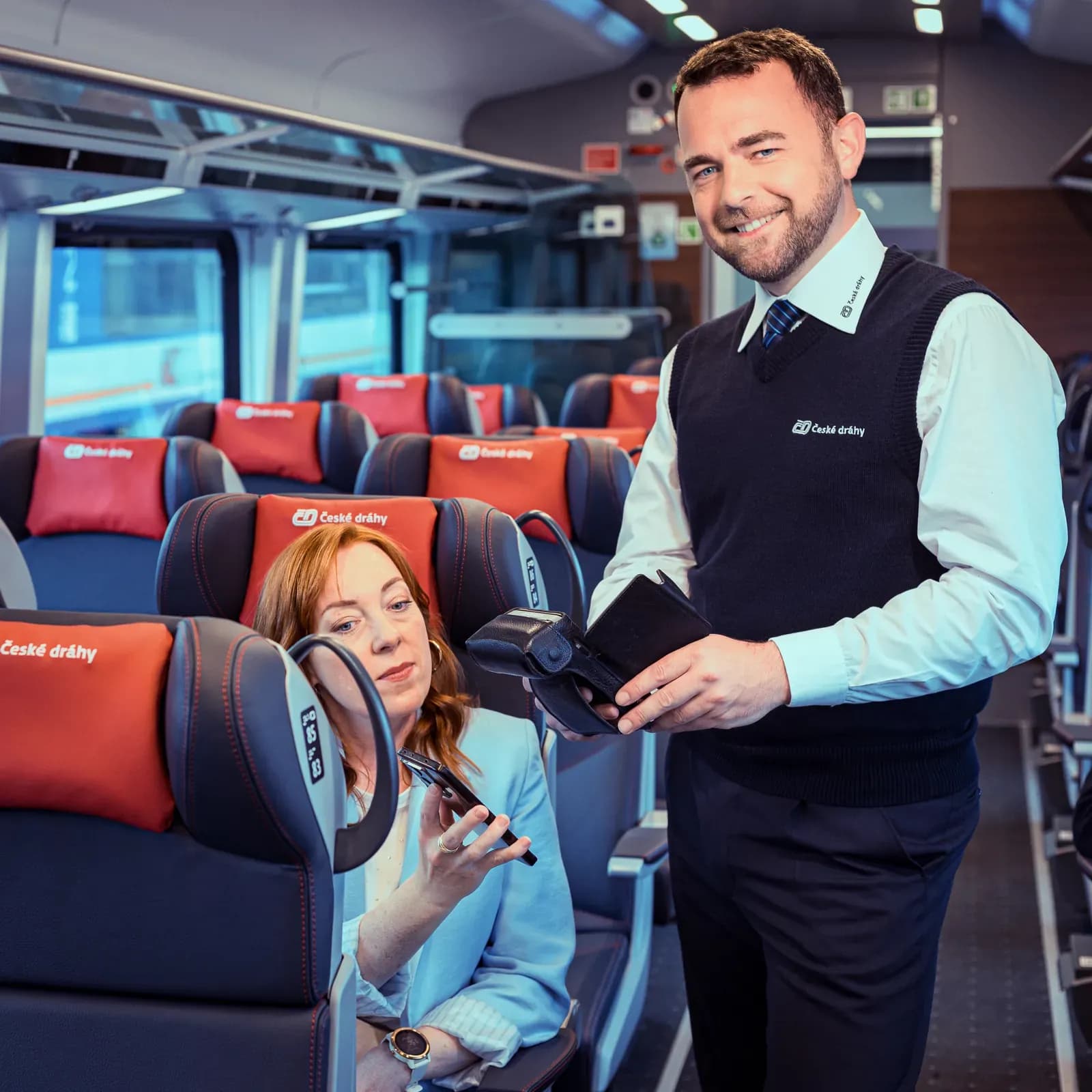 České dráhy conductor in a first class carriage holding a portable payment terminal while a seated passenger pays with her phone